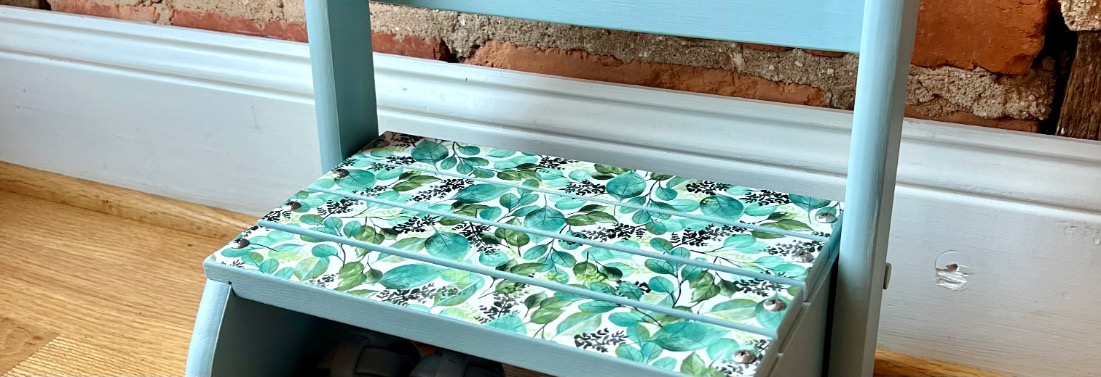 Stool-makeover-hero A small, light blue wooden step stool with a seat decorated in a leafy green and white pattern, placed on a wooden floor near a white baseboard and exposed brick wall.