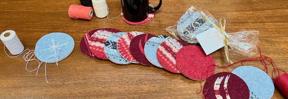Many colorful, cozy coasters made from old sweaters are seen laying on a table with spools of wool yarn being used to finish the edges with a blanket stitch