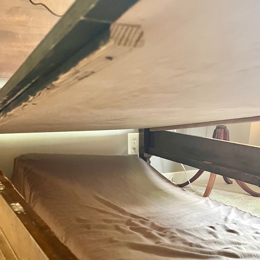 A low view under a bed shows an electrical outlet on the wall, a brown sheet covering part of the floor, and wooden furniture legs in the background—ideal for spaces fitting beds from Twin beds to X-Long.