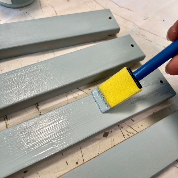 A hand uses a yellow foam brush to paint a light blue coat onto wooden slats, which are laid out on a surface covered with a white sheet.