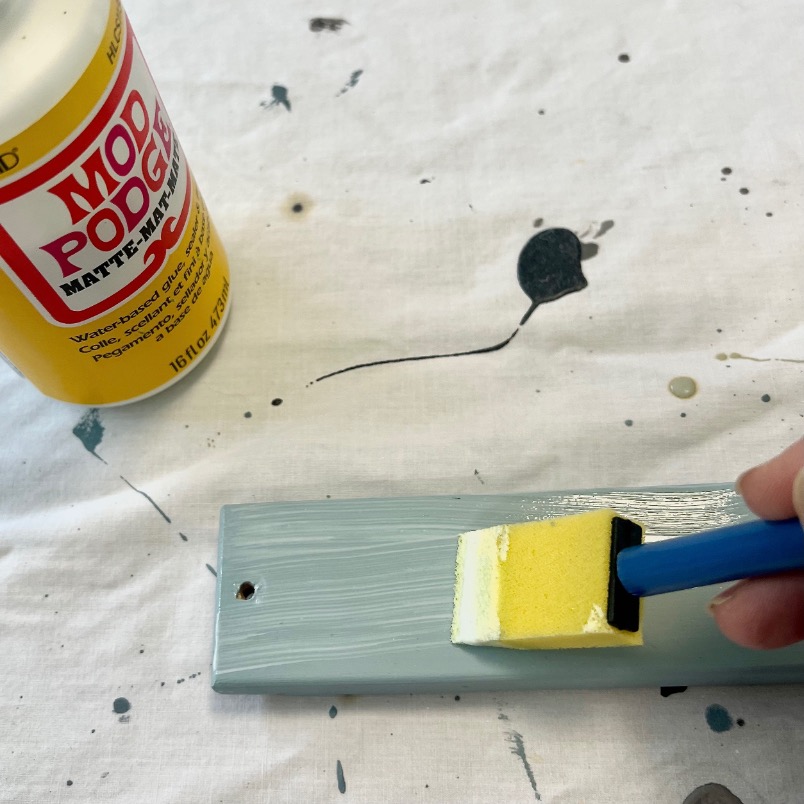 A hand uses a yellow foam brush to apply Mod Podge matte finish to a painted wooden piece on a white cloth with black paint splatters. The Mod Podge bottle is open and placed nearby.