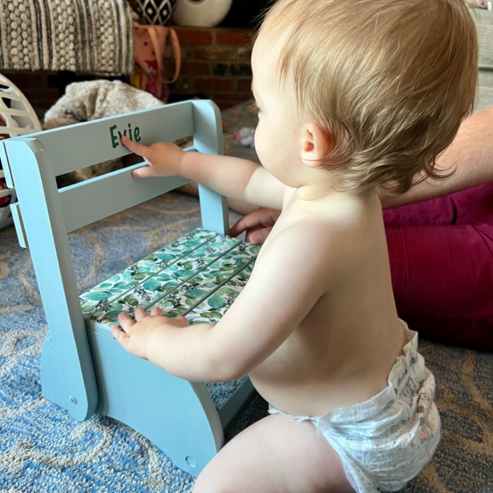 A baby in a diaper kneels on a blue carpet, playing with a small blue wooden chair labeled "Evie." An adult's arm is nearby, and toys and household items are in the background.