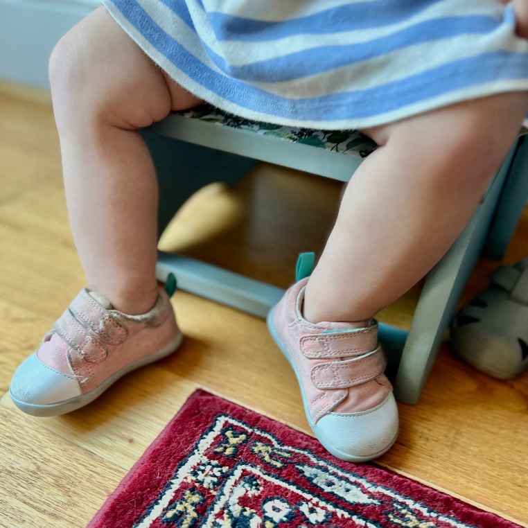 A small child wearing a blue and white striped dress sits on a chair, showing only their legs and feet in pink sneakers on a wooden floor near a patterned red rug.