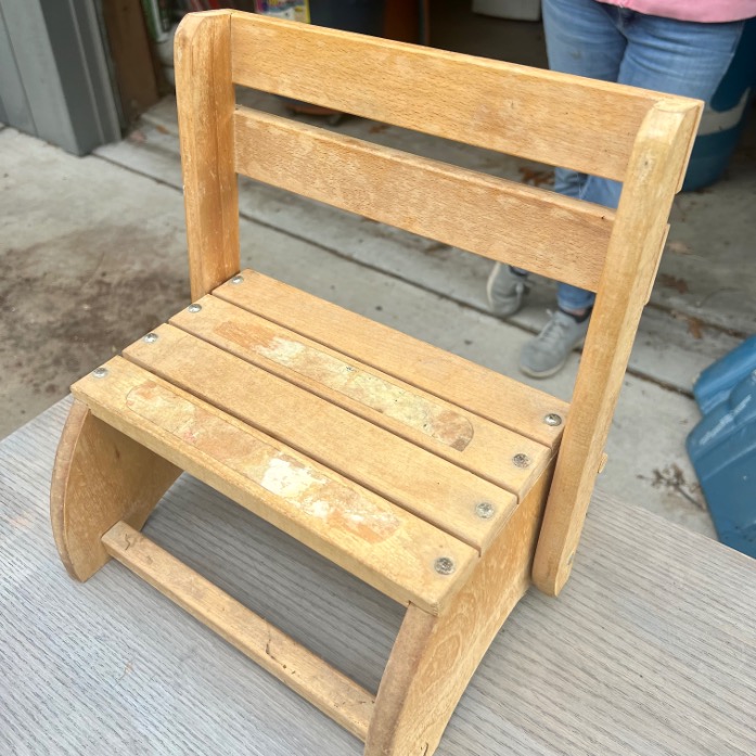A small wooden chair with a slatted seat and backrest sits on a table outdoors. The chair has a simple, rustic design and visible screws. A person wearing gray shoes and jeans stands in the background.