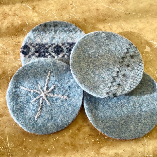Four round blue felt coasters, upcycled from an old sweater, feature simple stitched patterns like geometric and snowflake designs. They are arranged overlapping each other on a beige marbled surface.