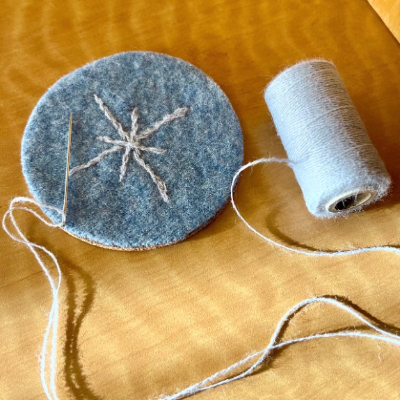 A round piece of blue felt, upcycled from an old sweater, with a white star-like embroidery pattern sits beside a spool of light blue thread and a sewing needle on a wooden surface.