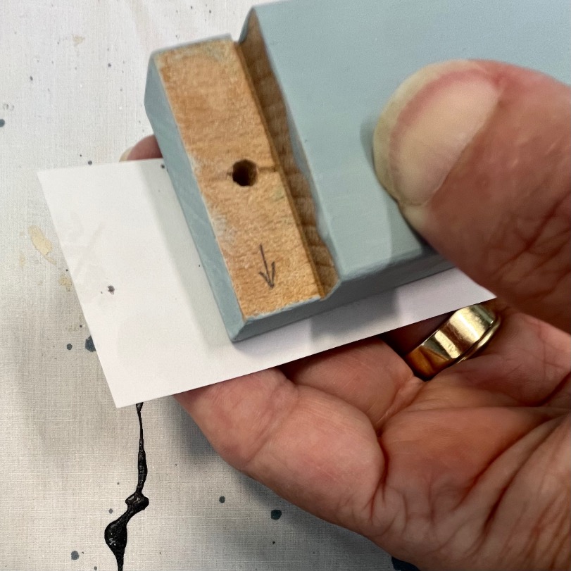 A hand holds a painted wooden block with a hole and an arrow drawn on its end, pressing it against a white piece of paper. The person is wearing a gold ring.