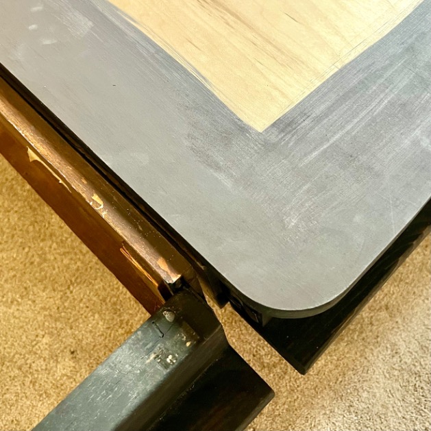 Close-up of a table corner with a metal frame, showing a smooth, rounded black edge and a light wood tabletop—perfect for rooms with twin beds to X-Long. The angled leg rests on beige carpet.