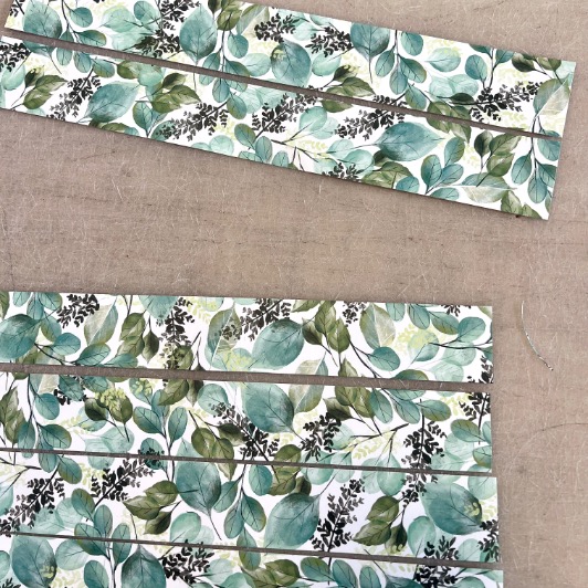 Rectangular strips of paper with a green leafy pattern are laid out on a neutral fabric surface. Some strips are grouped together at the bottom, while one strip is separated above.