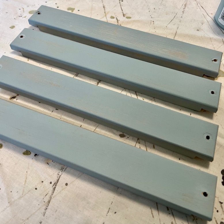 Four rectangular wooden boards painted light blue are laid side by side on a light-colored, speckled surface. Each board has a hole near one end.