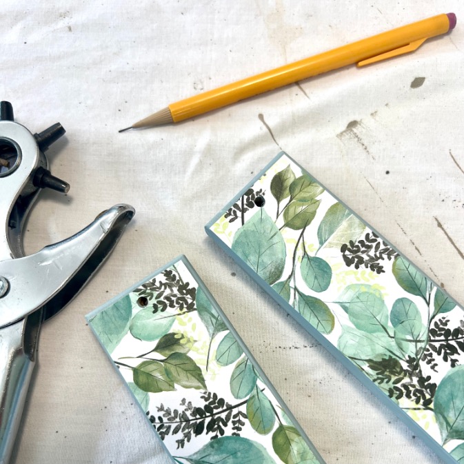 A yellow pencil, a metal hole punch, and two paper bookmarks with green leaf designs rest on a white, slightly stained fabric surface.