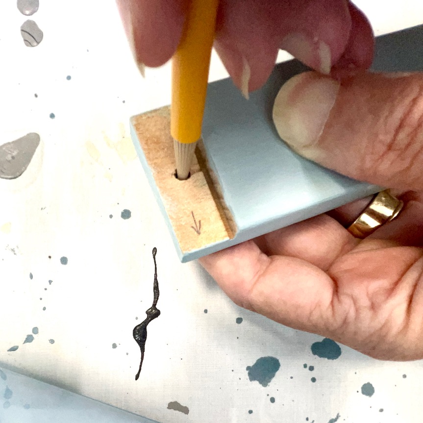 A hand holding a pencil marks a hole in the end of a light blue wooden piece. The person wears a gold ring, and the surface below is splattered with black and gray paint.