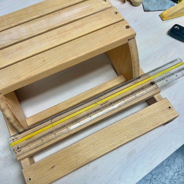 A small wooden stool lies on its side on a workbench with a clear ruler placed along one of its slats. Tools and materials are visible in the background.