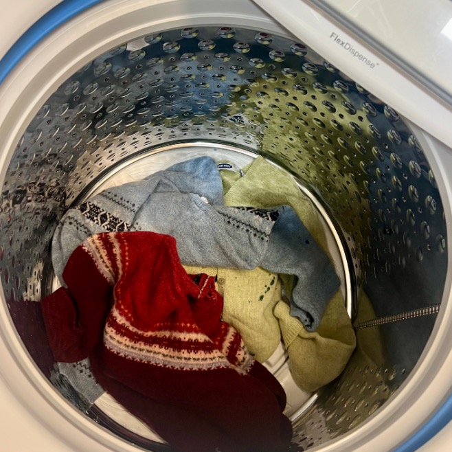 A top-down view of a washing machine drum shows a few colorful sweaters, including red, blue, and yellow garments—perfect inspiration if you want to upcycle old sweater pieces. The washer's metallic interior gleams in contrast.