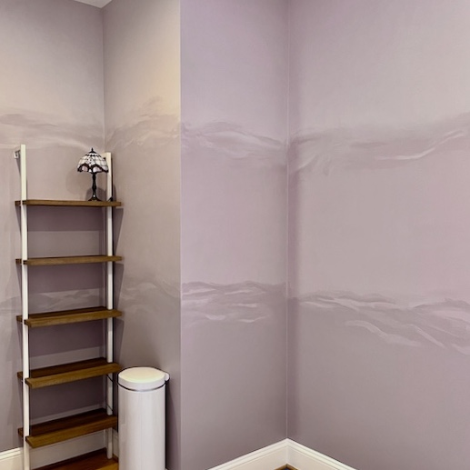 A corner of a room with pale purple walls, a five-tier wooden shelf with white supports, a small lamp on the top shelf, a diy-stool-makeover tucked nearby, and a white cylindrical trash can on the floor.