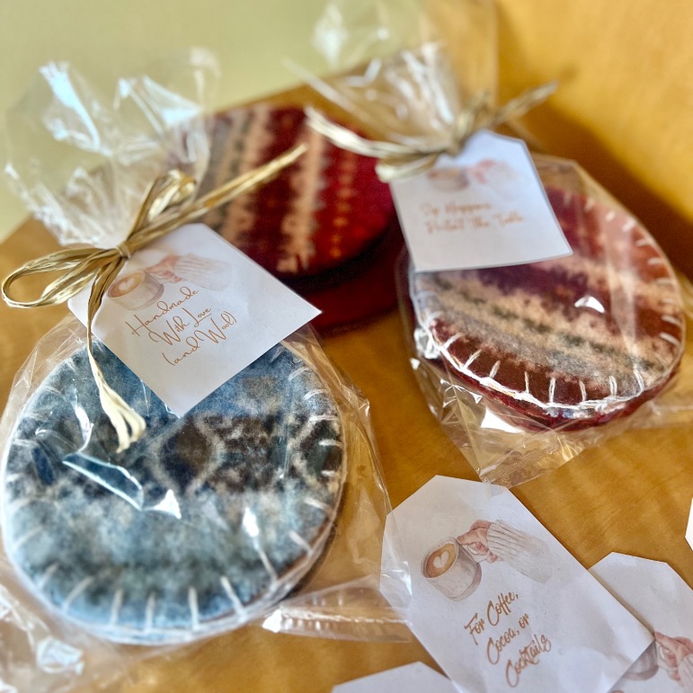 Handmade felt coasters, upcycled from an old sweater, in blue and red patterns are wrapped in clear plastic with gold ribbons and tags, displayed on a wooden surface with extra tags beside them.