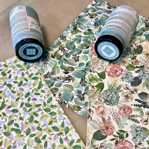 Two bottles of acrylic paint sit on top of three sheets of floral-patterned paper, each featuring different designs with leaves and flowers in various colors.