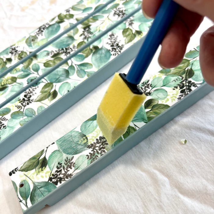 A hand uses a yellow foam brush to apply a clear coating over a wooden piece decorated with a leafy green and blue botanical pattern. Other similarly decorated wood pieces are visible in the background.