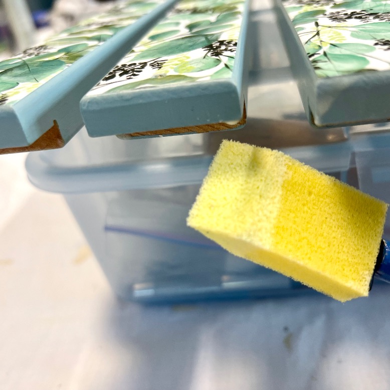 A yellow foam brush applies paint to the edge of a decorative tile with a floral pattern; other similar tiles are visible, drying above a plastic container.