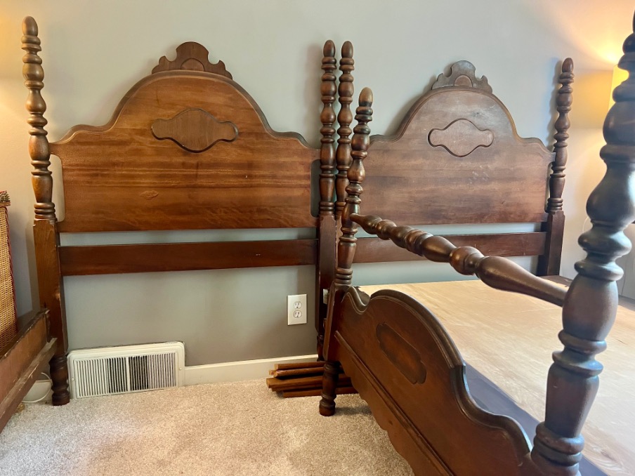 Two vintage wooden twin bed frames, convertible from twin beds to X-Long, feature tall, turned posts and decorative headboards. They sit side by side in a carpeted room with gray walls, a vent, and an electrical outlet behind them.
