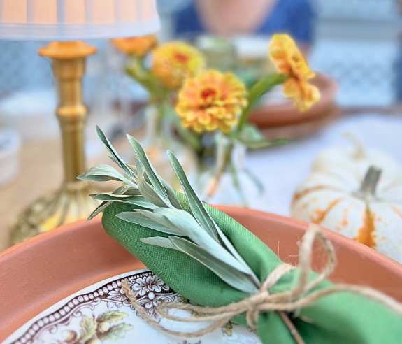 A close-up of a table setting with a terracotta plate, a green napkin tied with twine and greenery, an ornate plate, yellow flowers, a white pumpkin, and a gold lamp in the background.