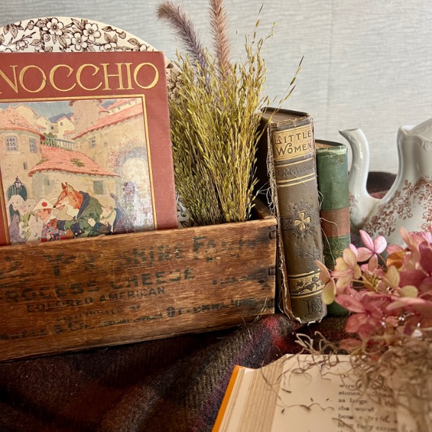 A rustic wooden box serves as a charming book centerpiece, holding a vintage "Pinocchio" book, dried grasses, and classic novels, arranged with a floral teapot and pink flowers on a plaid cloth. An open book lies in the foreground.