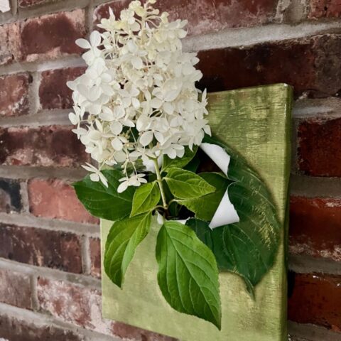 A cluster of white hydrangea flowers with green leaves is arranged in a small canvas pouch mounted on a green-painted square, hanging on a brick wall.