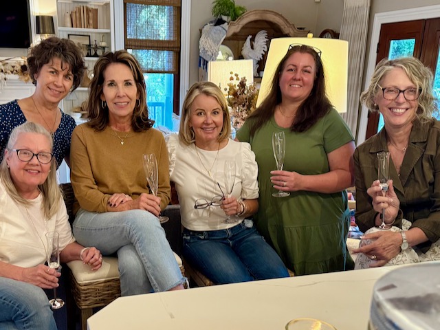 Six women sit together indoors, smiling at the camera and holding empty champagne glasses, suggesting a casual, celebratory gathering in a cozy, warmly lit living space.