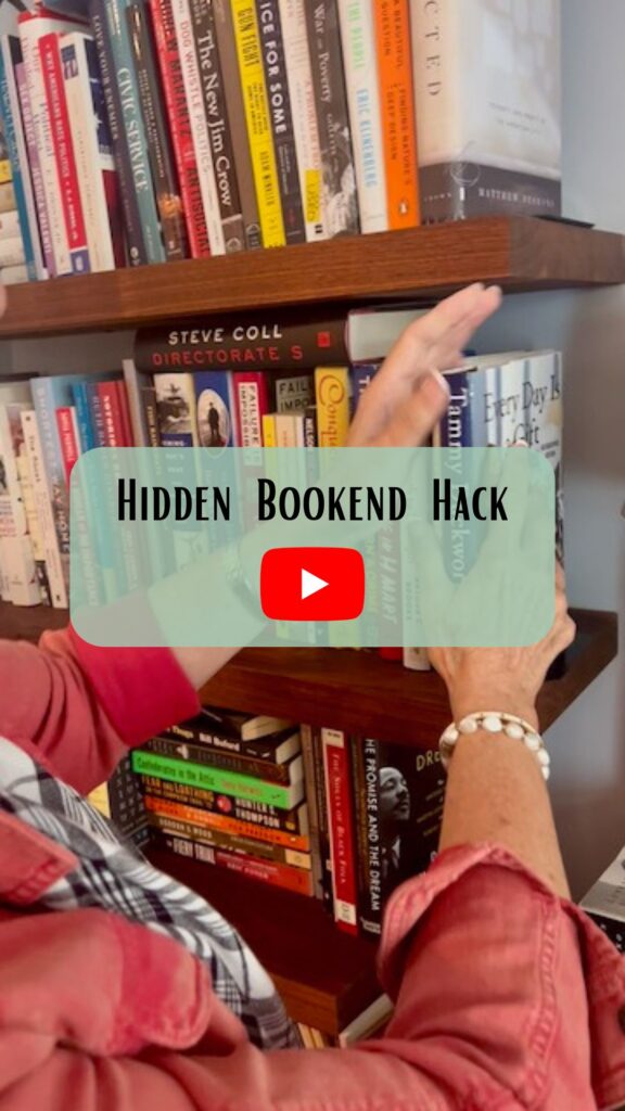 A person adjusts books on a shelf, concealing a metal bookend behind them. Overlaid text reads “Hidden Bookend Hack” with a red play button symbol, suggesting a video tutorial.
