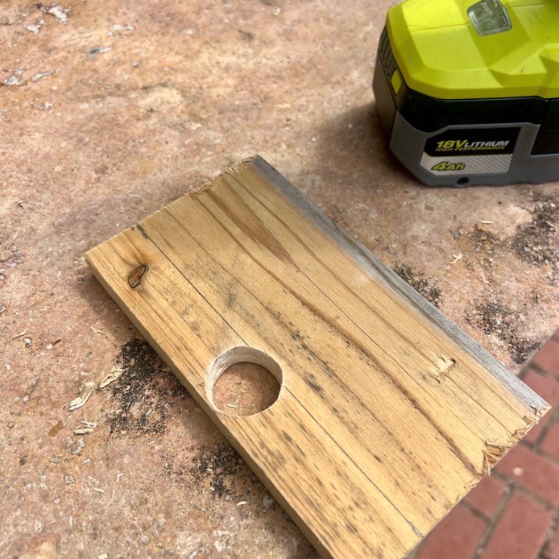 A piece of wood with a circular hole drilled through it rests on a work surface. Nearby is a green and black rechargeable power tool battery. Wood shavings are scattered around.