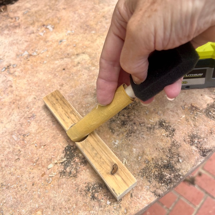 A hand uses a foam brush to apply a finish or stain to a small wooden object on a brown, speckled surface outdoors.