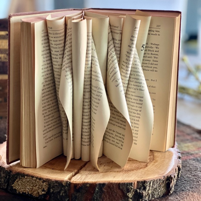 An open book with its pages creatively folded inward forms a wavy, sculptural pattern—a stunning Book Centerpiece resting on a round wooden slab, with a blurred background.