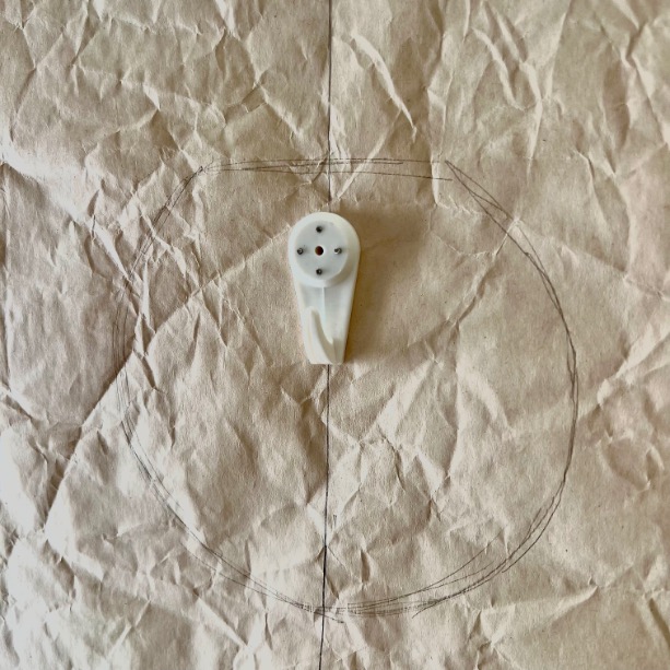 A small white plastic object with three holes, ideal to hang gallery wall pieces, rests at the center of a hand-drawn circle on crumpled brown paper.