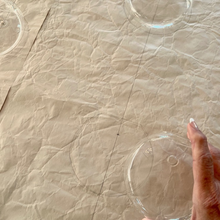 A hand with a French manicure traces a clear round object onto crumpled brown paper, creating visible circle outlines and straight pencil lines—perfect for planning to hang a gallery wall with precise alignment.