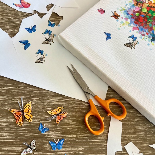 A pair of orange-handled scissors, cut-out paper butterflies, and a canvas with colorful butterfly art are on a wooden table, with scraps of white paper scattered around.