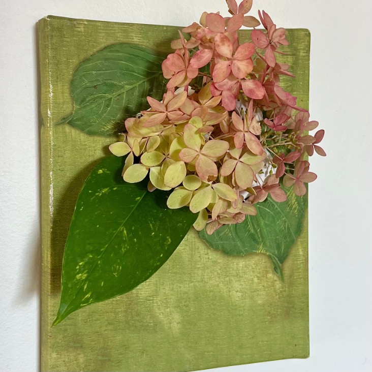 A mixed media artwork featuring pink and pale yellow hydrangea flowers and green leaves arranged on a textured, light green canvas and mounted on a white wall.