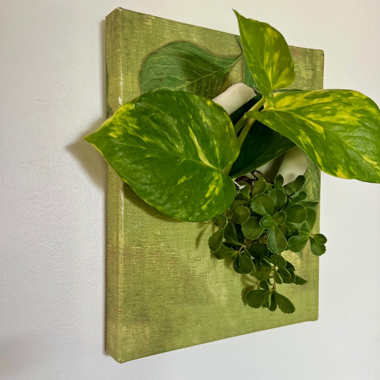 A wall-mounted green canvas holds several lush green plants, including a pothos and another leafy plant, creating a vertical garden art piece against a white wall.