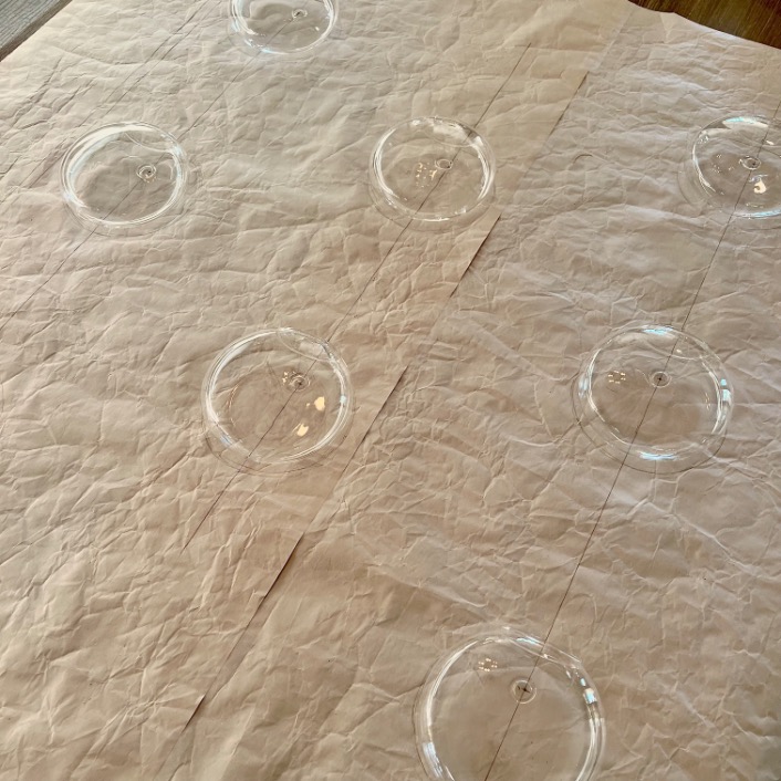 Clear plastic ornaments rest on crinkled sheets of white paper, evenly spaced in a grid pattern—like pieces on a gallery wall ready to hang. The well-lit surface highlights the texture of the paper and the ornaments' transparency.