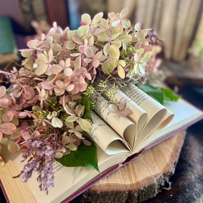 An open book lies on a wooden surface with its pages artistically folded into heart shapes, creating a charming Book Centerpiece that holds a bouquet of pastel pink and purple flowers alongside lush green leaves.