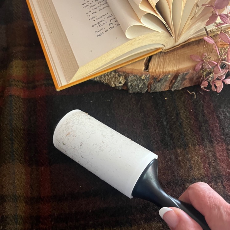 A hand holding a lint roller with debris on it, next to a book centerpiece and dried flowers on a wooden surface, all arranged on dark plaid fabric.