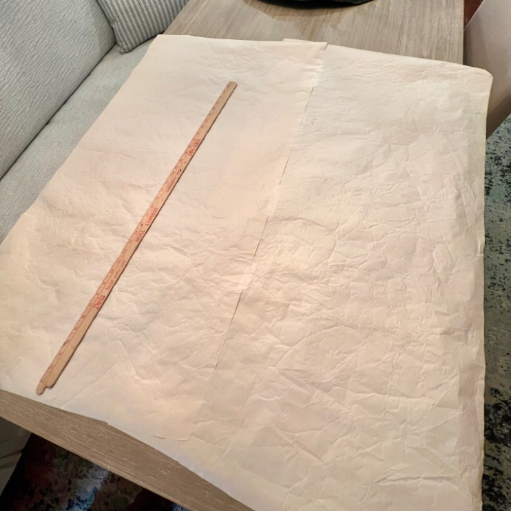 Two large sheets of wrinkled parchment paper lie on a wooden surface next to a sofa, with a wooden yardstick placed diagonally across the papers—perfect for planning your hang gallery wall layout.