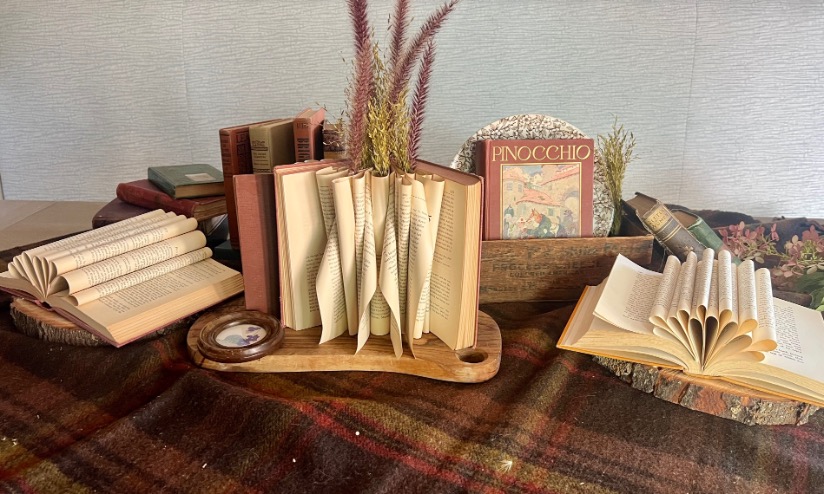 Books with pages artistically folded into shapes create a charming book centerpiece, displayed on wooden trays and a plaid cloth, with dried grasses and a vintage copy of "Pinocchio" in the background.