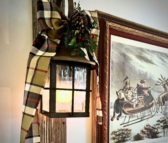 A lantern with a lit candle hangs on a wood beam, decorated with a plaid ribbon, pinecones, and greenery. Beside it is a framed winter painting of people riding in a horse-drawn sleigh through the snow.