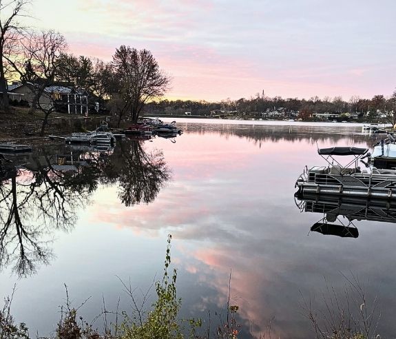 A calm lake at sunset with pink clouds reflected in the water, boats docked along the shore, houses and bare trees lining the left side, and soft light illuminating the scene.