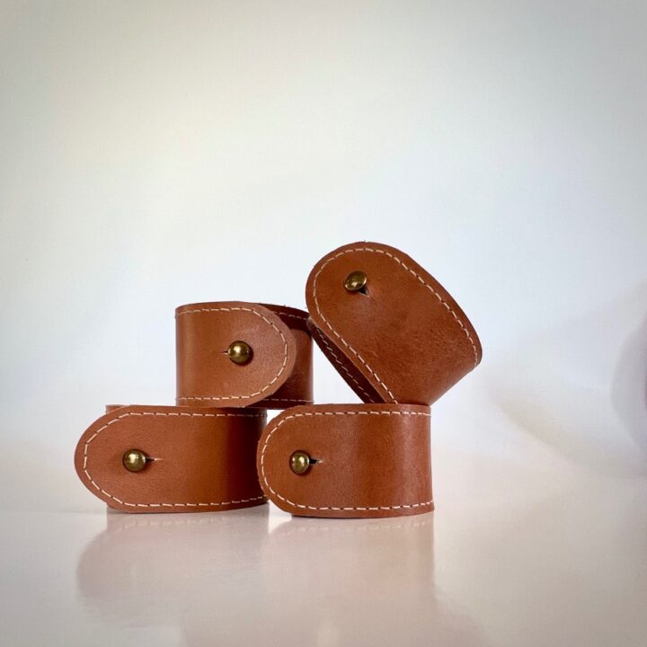 Four brown DIY leather napkin rings with white stitching and brass rivets are stacked together on a white surface, set against a plain white background.