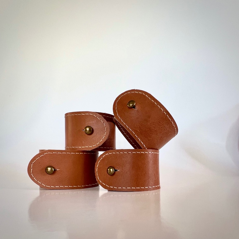 Four brown DIY leather napkin rings with white stitching and brass rivets are stacked together on a white surface, set against a plain white background.