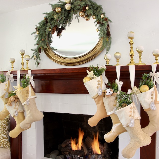 A beautifully decorated christmas mantel showcases a round mirror, gold candlesticks, a green garland, and six cream-colored Christmas stockings filled with gifts hanging above the lit fireplace.