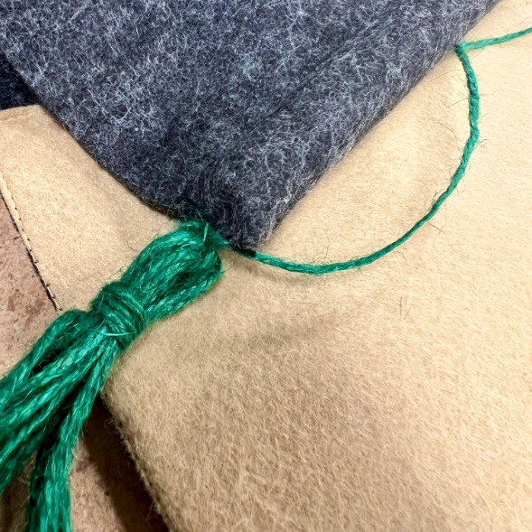 A close-up of layered felt sheets in tan and dark gray colors, stitched together with bright green yarn and a green yarn tassel resting on top.