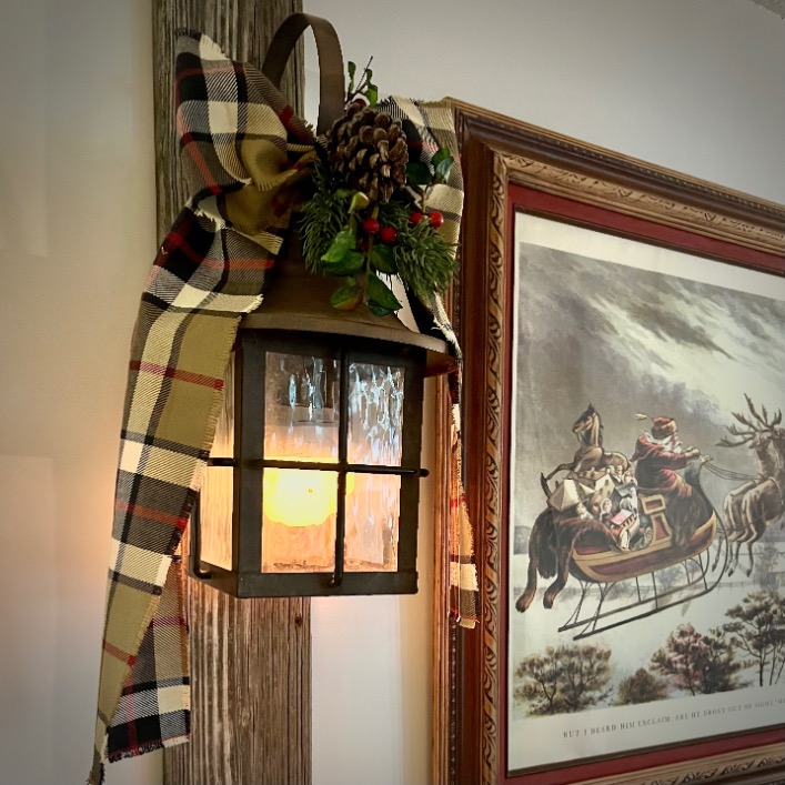 A rustic lantern with a lit candle hangs on a wooden beam, decorated with a plaid ribbon, pinecone, and greenery—a scene inspired by Ralph Lauren Christmas ideas. Nearby, a framed artwork shows Santa Claus riding a sleigh through the snow.