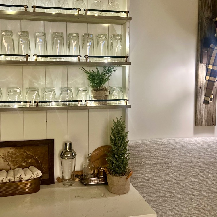 Shelves lined with upside-down glasses are mounted on a wall above a countertop. The counter, inspired by Ralph Lauren Christmas ideas, holds potted plants, a cocktail shaker, rolled napkins in a basket, and small decor items for a cozy bar nook.